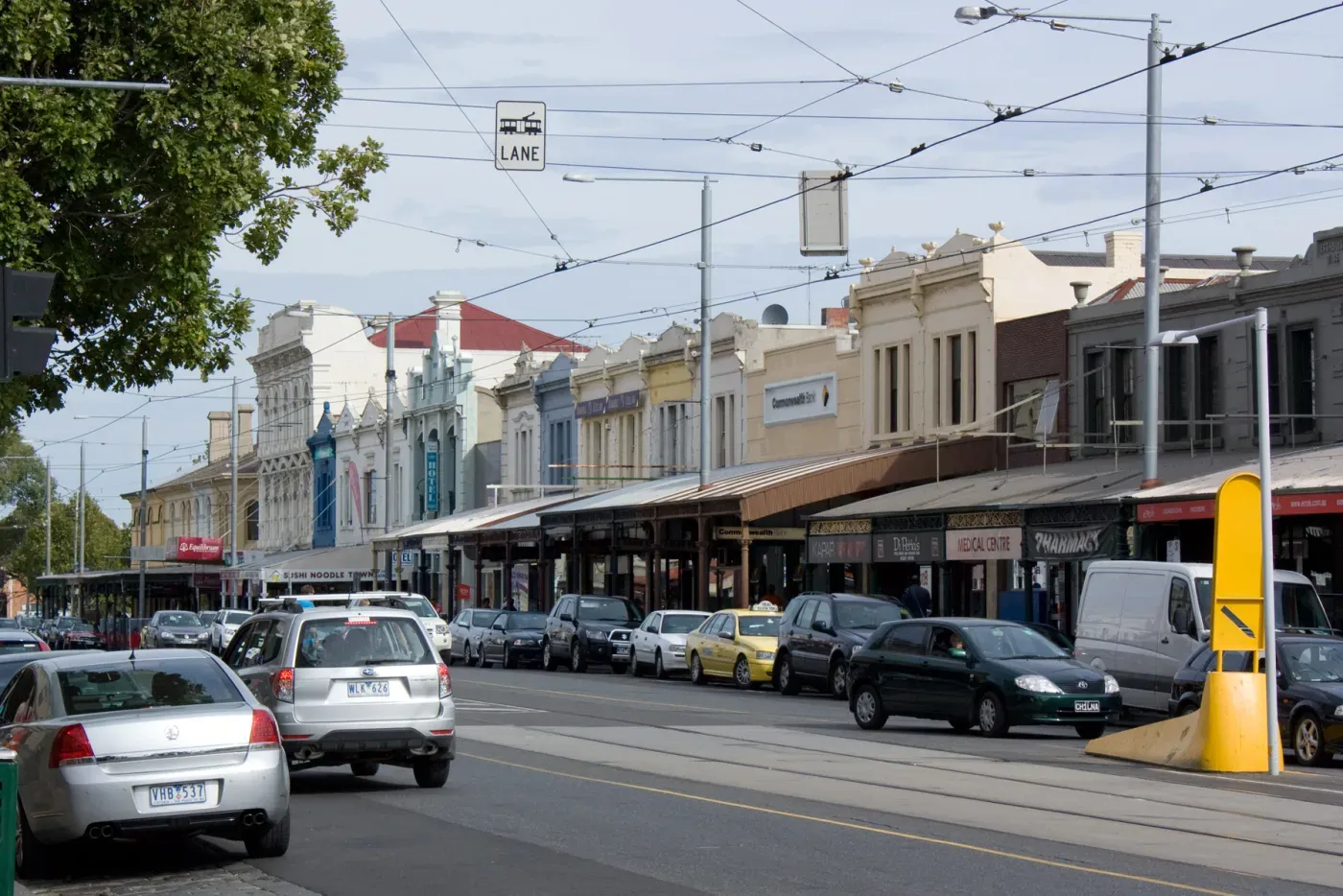 An inner-suburban shopping strip on Errol Street, North Melbourne, with heritage shopfronts, awnings, tram tracks and parked cars along the road.