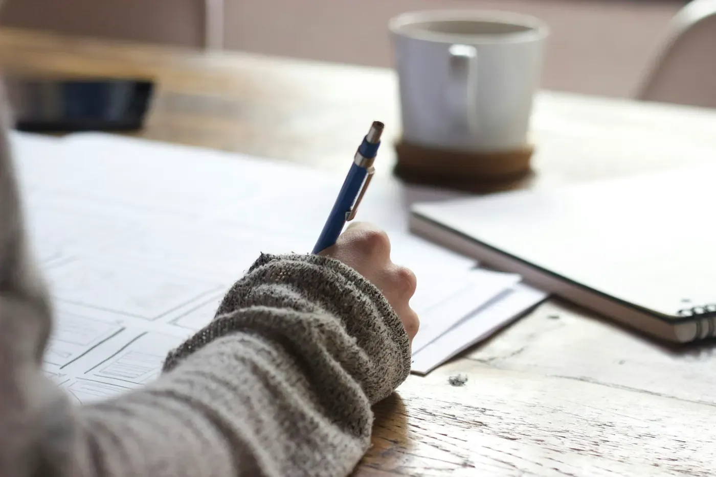 Hand taking notes beside a laptop and a coffee mug.
