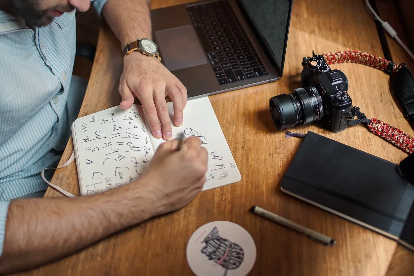Looking down at a wooden desk where a person in a striped shirt sketches handwritten lettering in an open notebook, beside a laptop, a camera, a closed black notebook and a pen.
