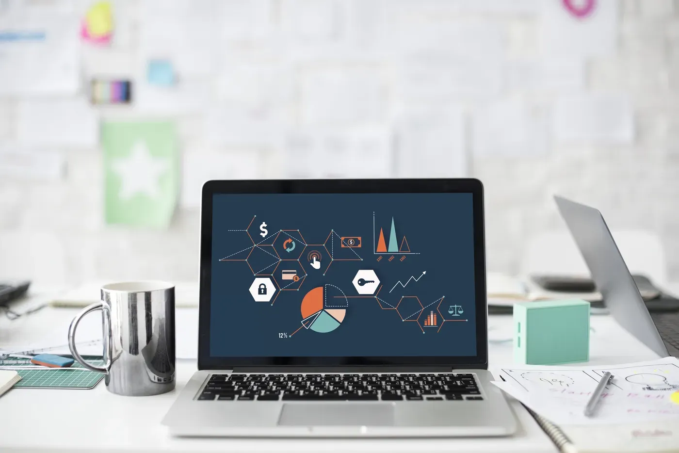 An open laptop on a white desk shows an illustration of linked data icons, including a padlock, a key, a pie chart and scales, beside a metal mug and sketched planning notes.