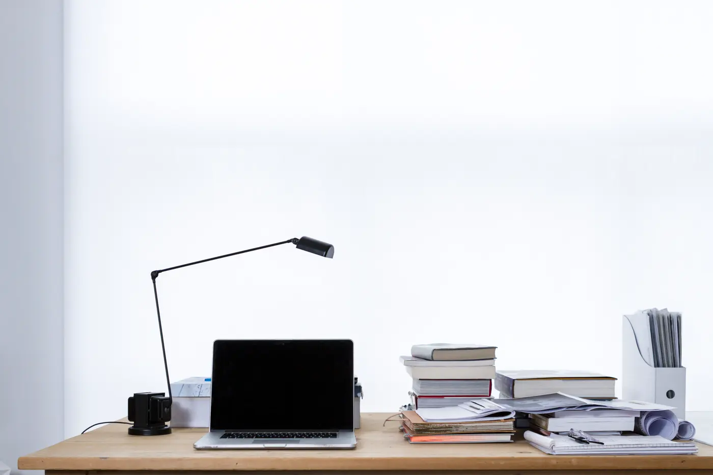 A closed-screen laptop and an angled black desk lamp on a wooden desk, beside stacks of books, folders and papers against a bright white wall.