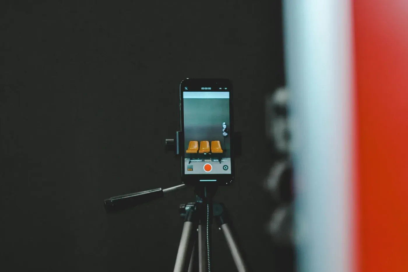 A smartphone mounted on a tripod recording video in a dark studio, its screen showing a camera app in record mode.
