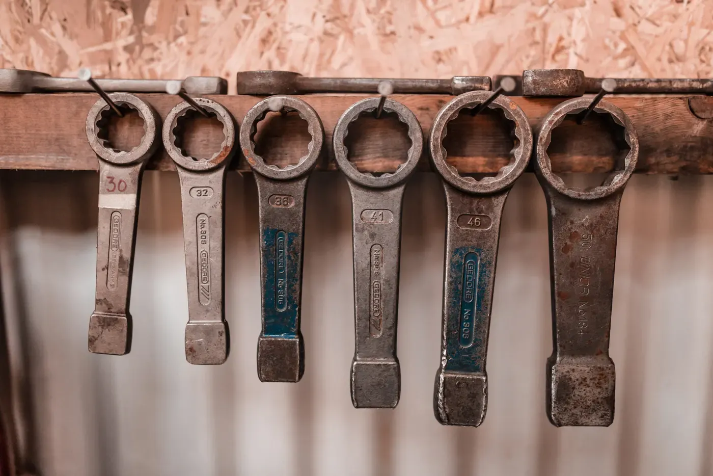 Six worn ring spanners hanging in size order from nails on a wooden rail, each stamped with its size from 30 to 50, against a pale workshop wall.