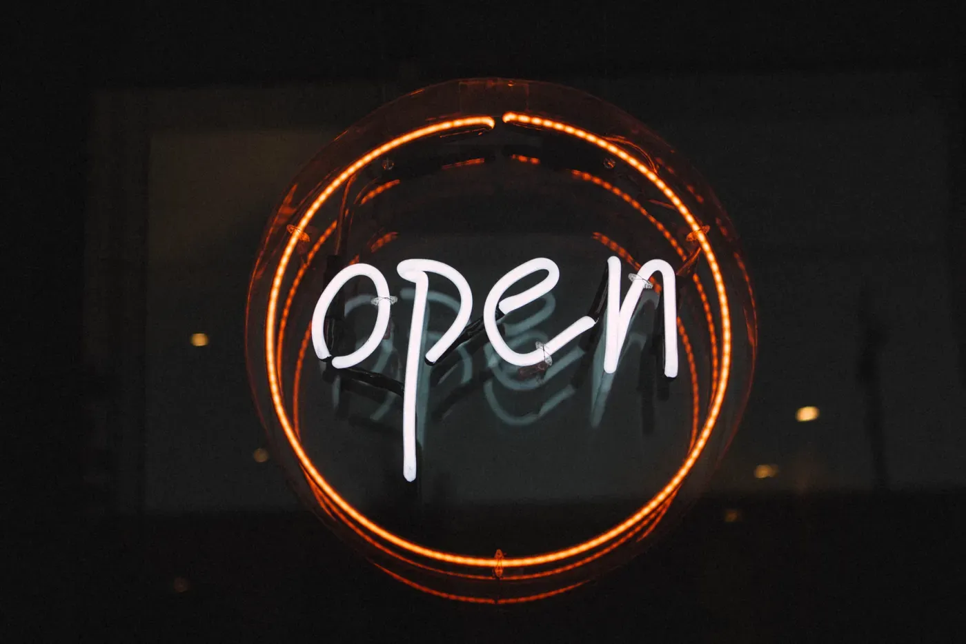 A round neon sign spelling "open" in white script inside a glowing orange ring, hanging in a dark shop window at night.