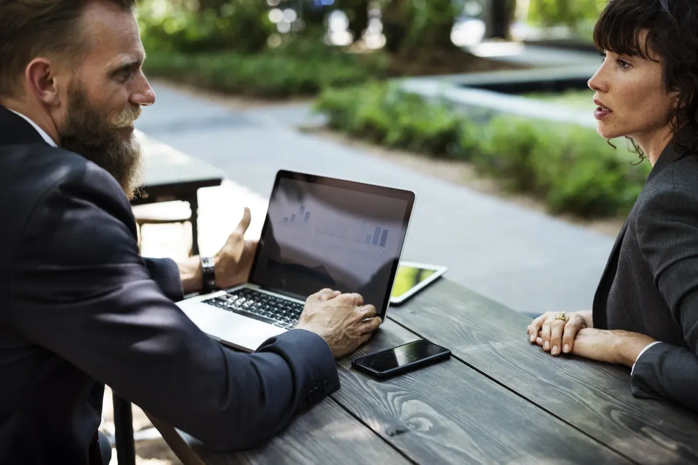 Two people in business attire talk across an outdoor wooden table, with an open laptop showing a bar chart, a tablet and a phone between them.