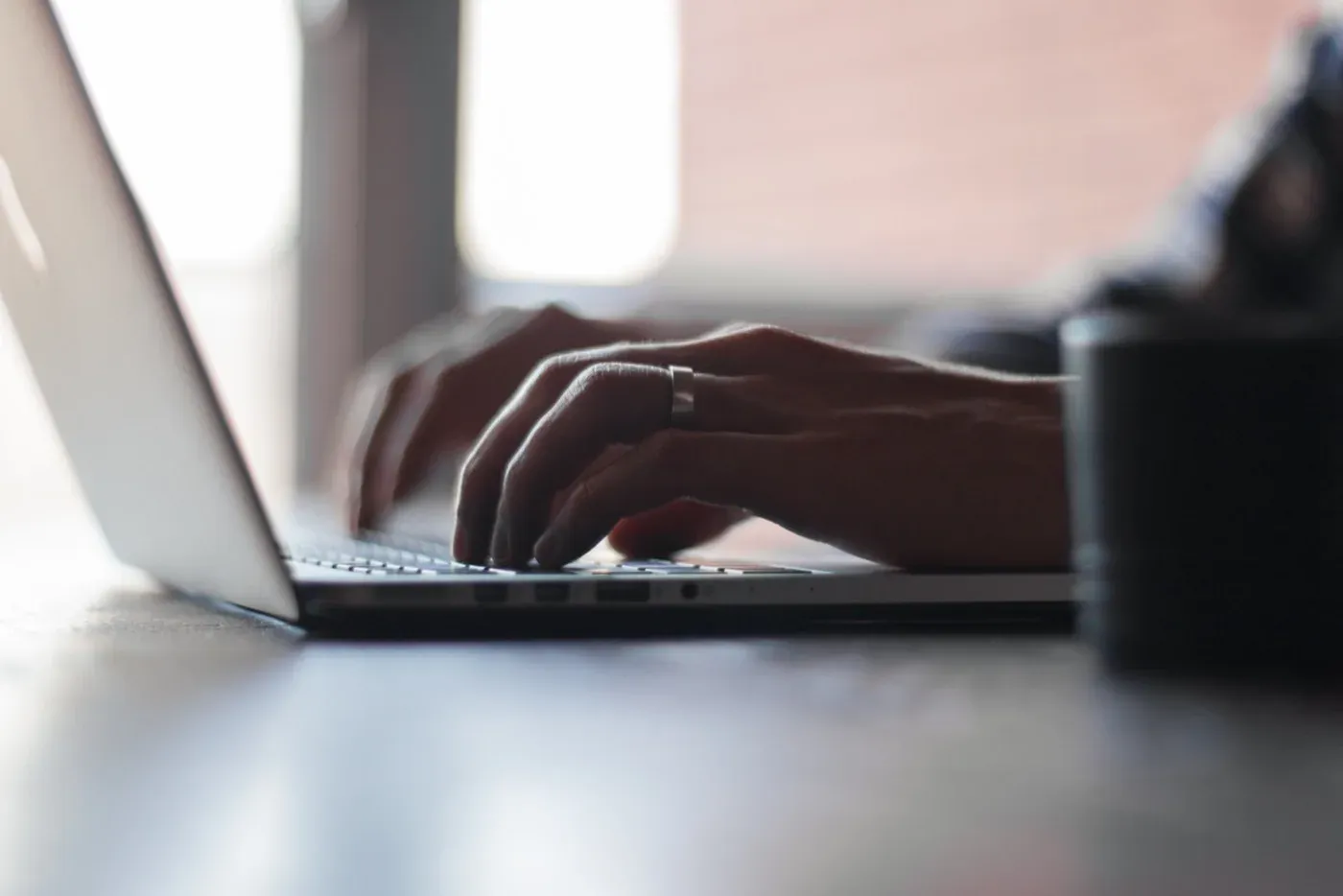 Close-up of a person's hands typing on a laptop keyboard on a wooden desk, with a blurred coffee cup and soft daylight behind.