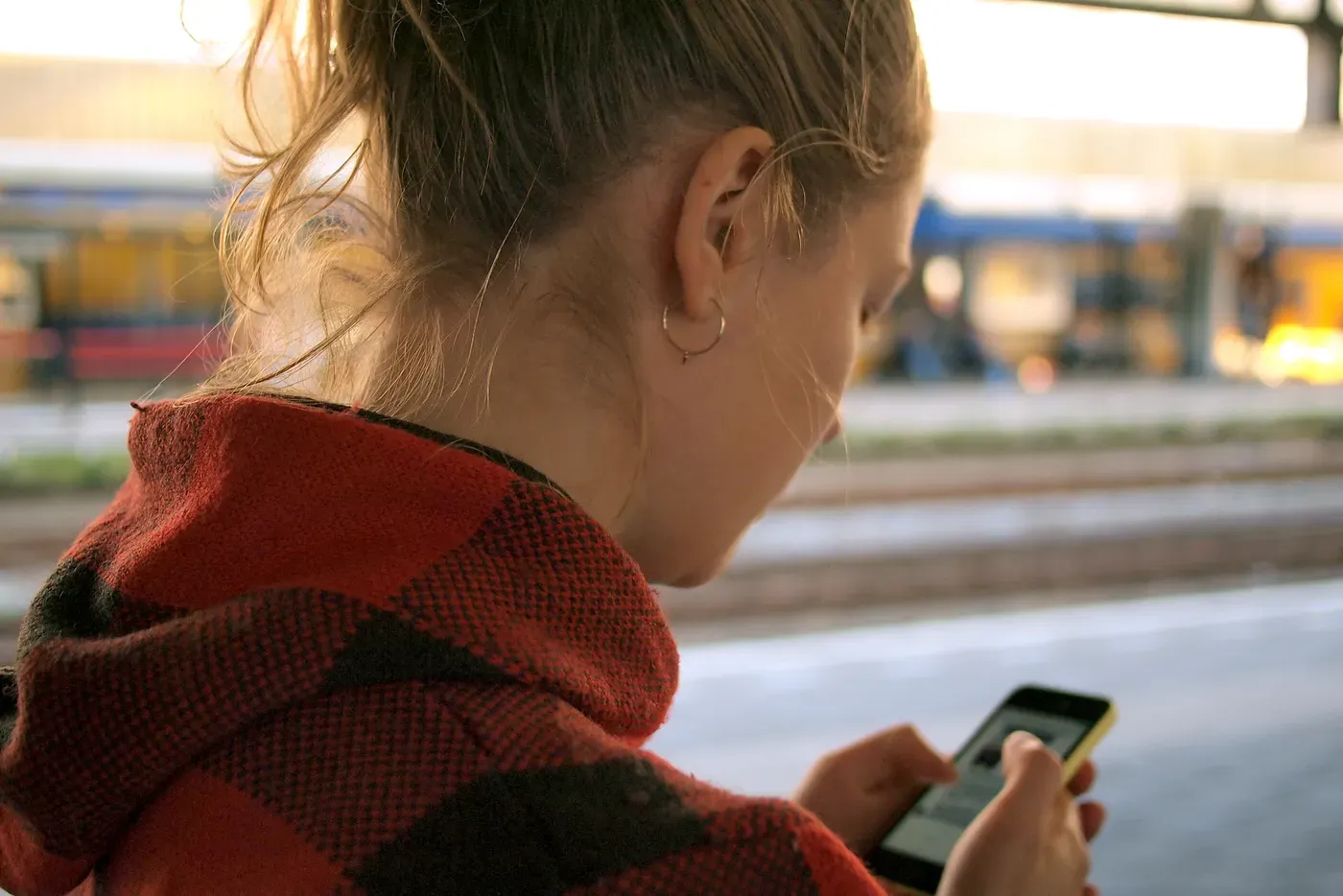 Seen from over her shoulder, a woman in a red and black checked hooded top looks down at a mobile phone in her hands on a train station platform, with a blurred train behind her.