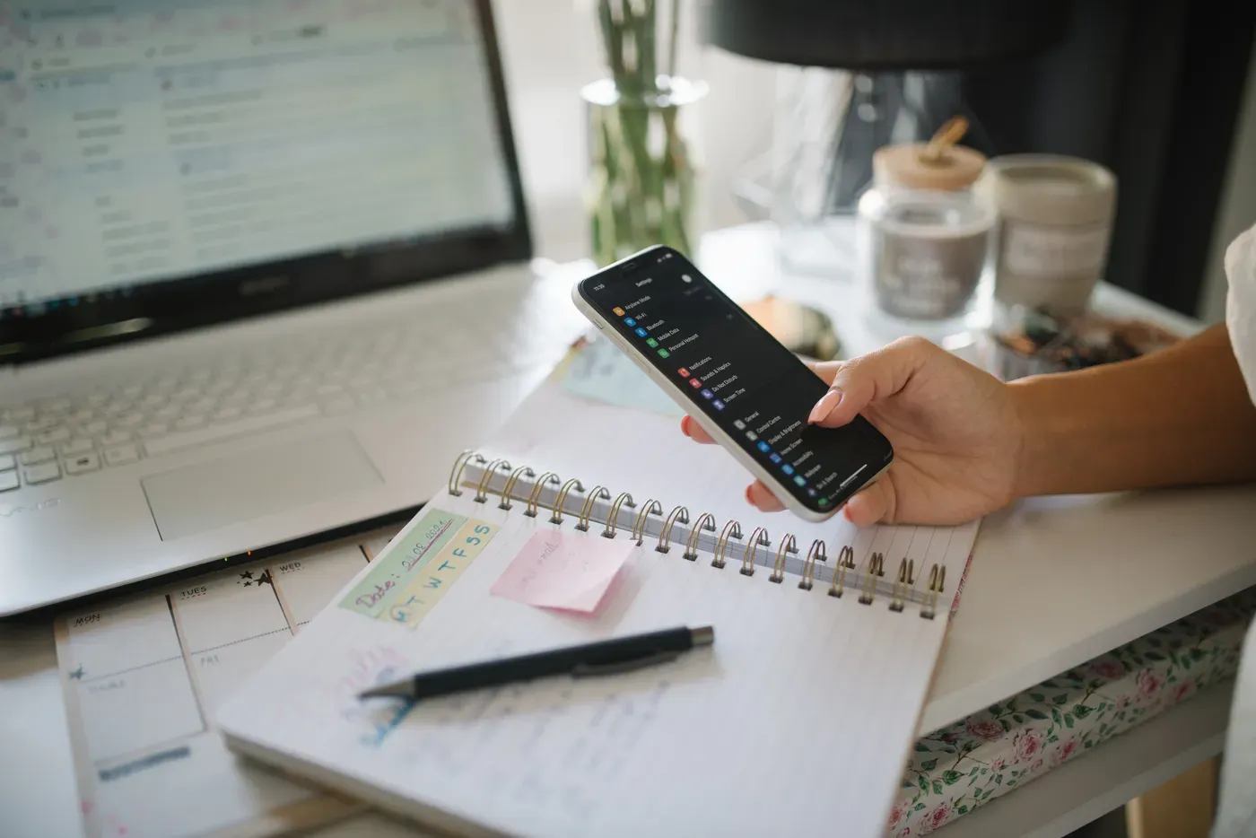 A woman's hand holds a smartphone over an open spiral notebook and a pen, with a laptop showing a blurred email inbox in the background.