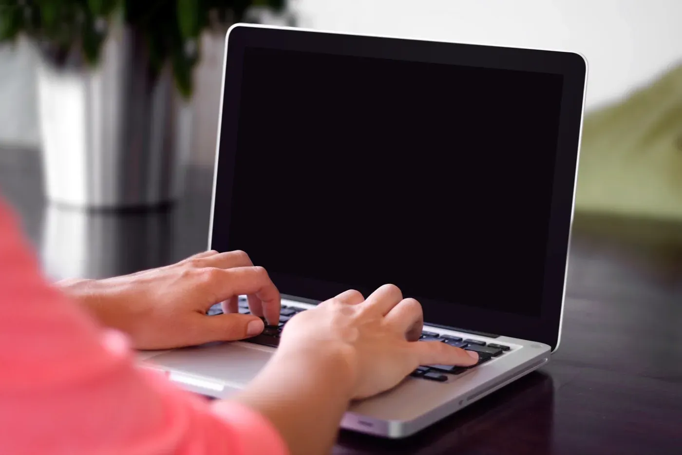 A person's hands typing on a laptop with a blank screen at a desk, with a pot plant blurred in the background.