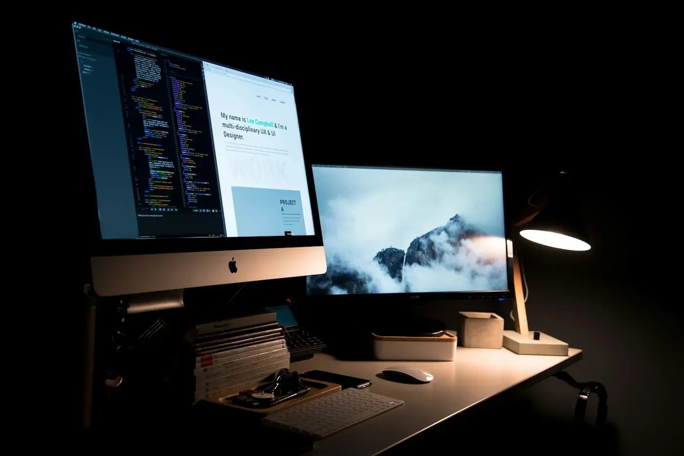Two monitors showing a website and code in a softly lit workspace.
