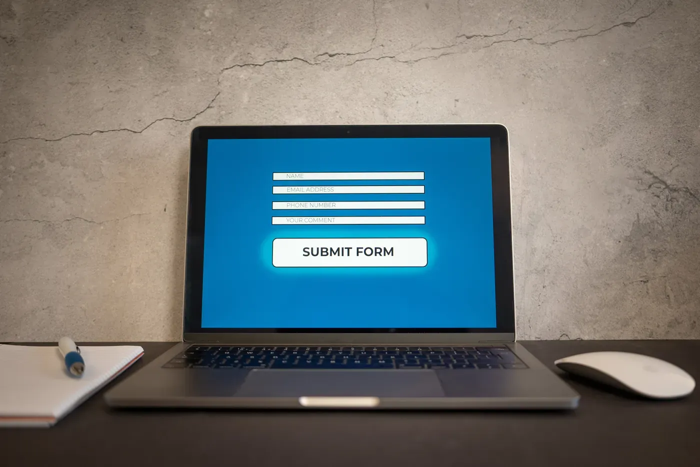 An open laptop on a dark desk against a concrete wall, its screen showing a simple blue contact form with name, email address, phone number and comment fields above a Submit Form button. A notepad and pen sit to the left and a white mouse to the right.