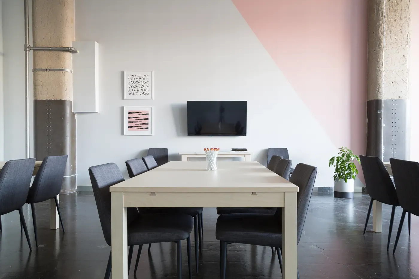 An empty meeting room with a long pale wooden table, grey chairs on both sides, a jar of pencils, a wall-mounted screen and a pink and white painted wall.