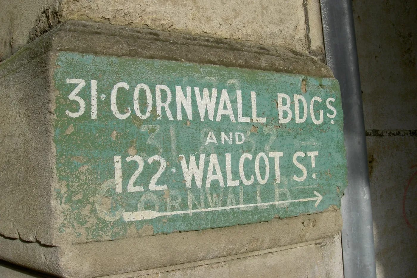 A weathered green painted sign on a stone building corner reads 31 Cornwall Bdgs and 122 Walcot St above a long white arrow, with faded lettering from an earlier version of the address still visible underneath.