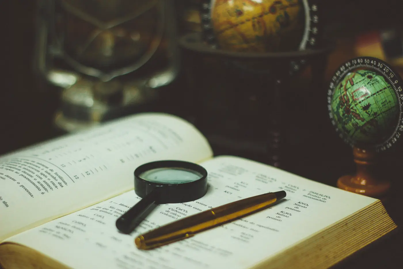 A black magnifying glass and a gold pen resting on an open printed book, with two small desk globes behind them in a dimly lit room.