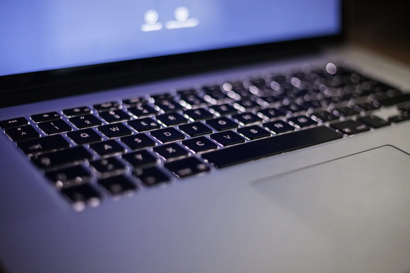 Close-up of a backlit laptop keyboard in a dim room, with the out-of-focus screen glowing blue behind it.