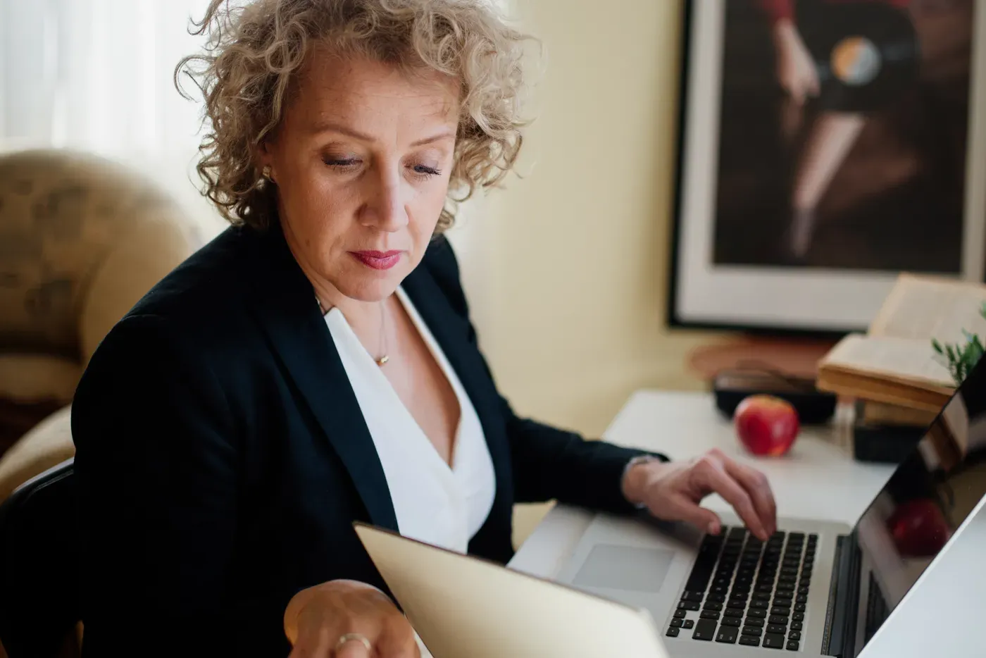 A woman with curly fair hair, wearing a dark blazer, sits at a desk turning the page of a notebook while she types on a laptop, with an apple and a stack of books behind it.