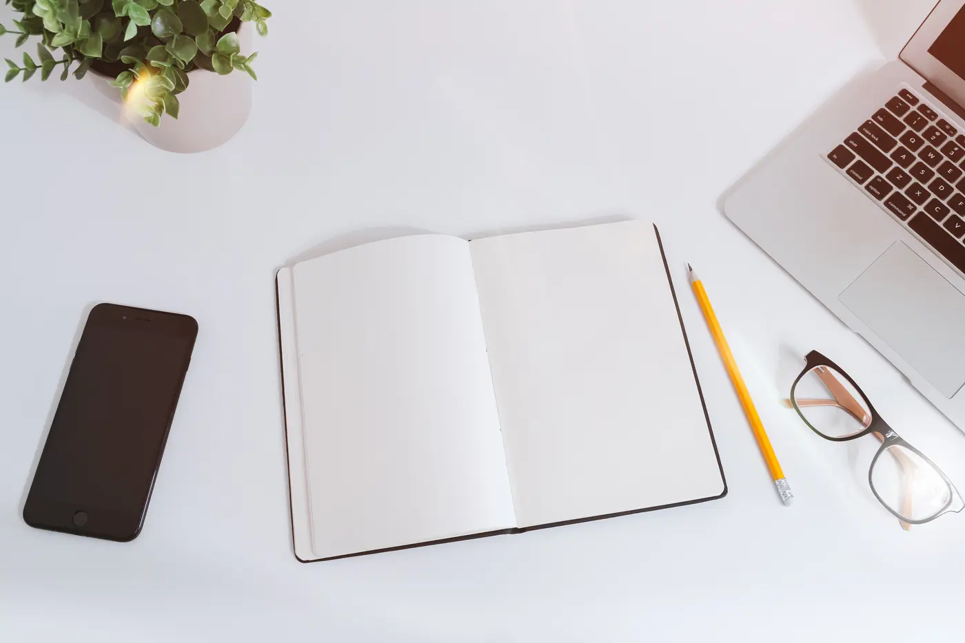 An open blank notebook and a yellow pencil on a white desk, with a smartphone, a pair of glasses, a laptop keyboard and a potted plant around it, seen from above.