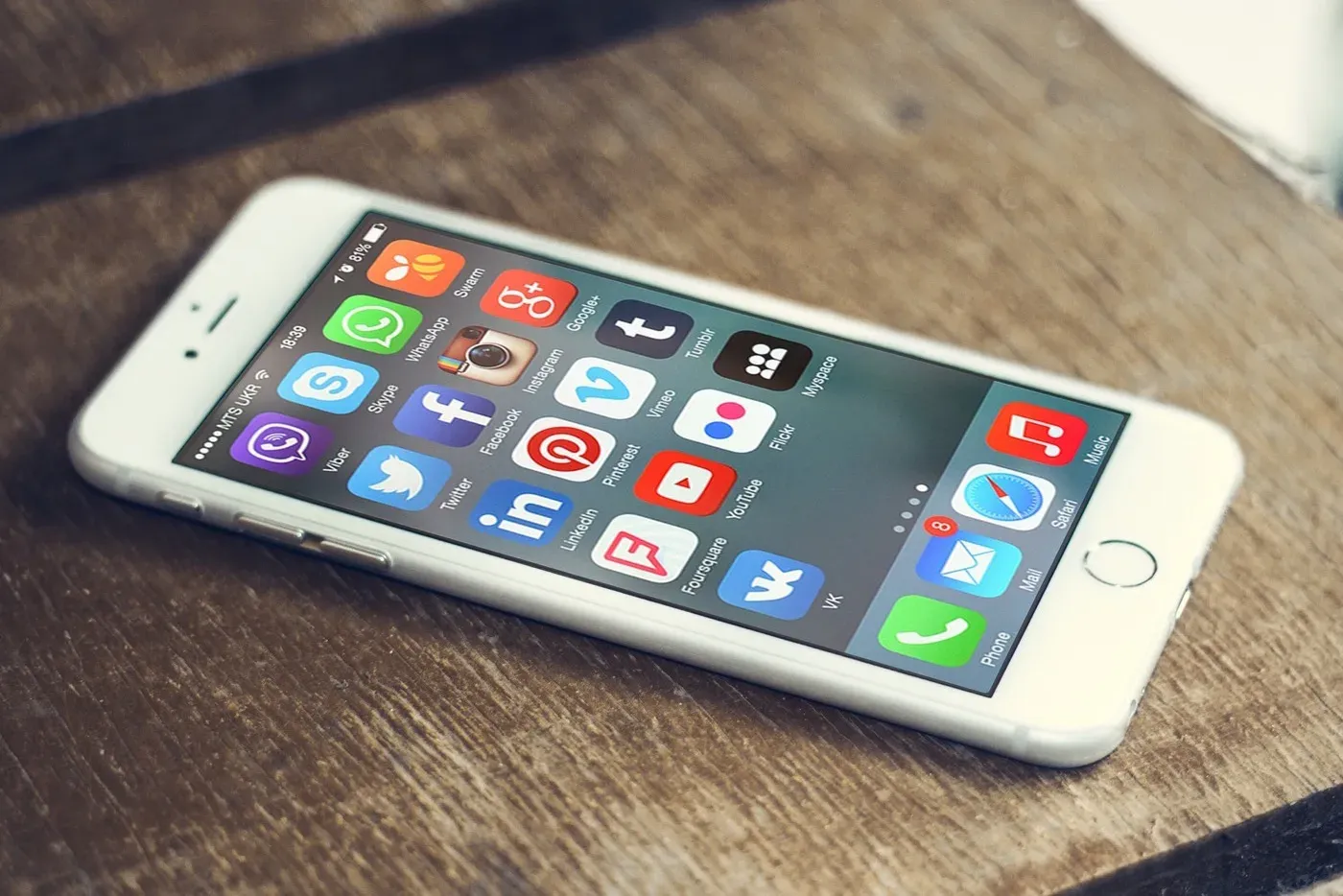 A white smartphone resting on a wooden desk, its screen showing a grid of colourful social media and messaging app icons.