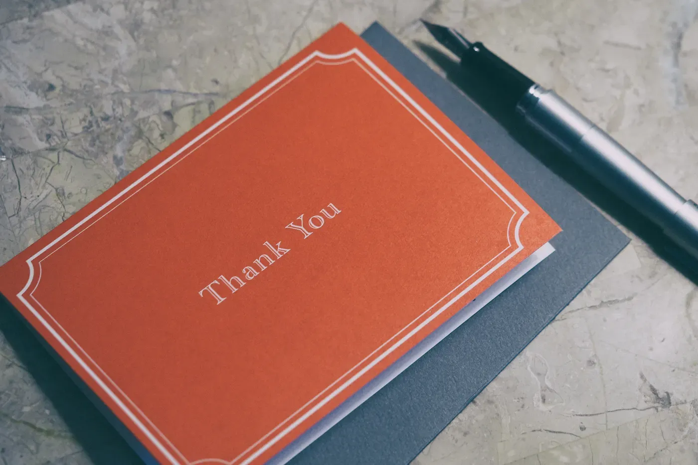 A red card printed with the words Thank You and a white double border, lying on a grey envelope beside a silver fountain pen on a marble surface.