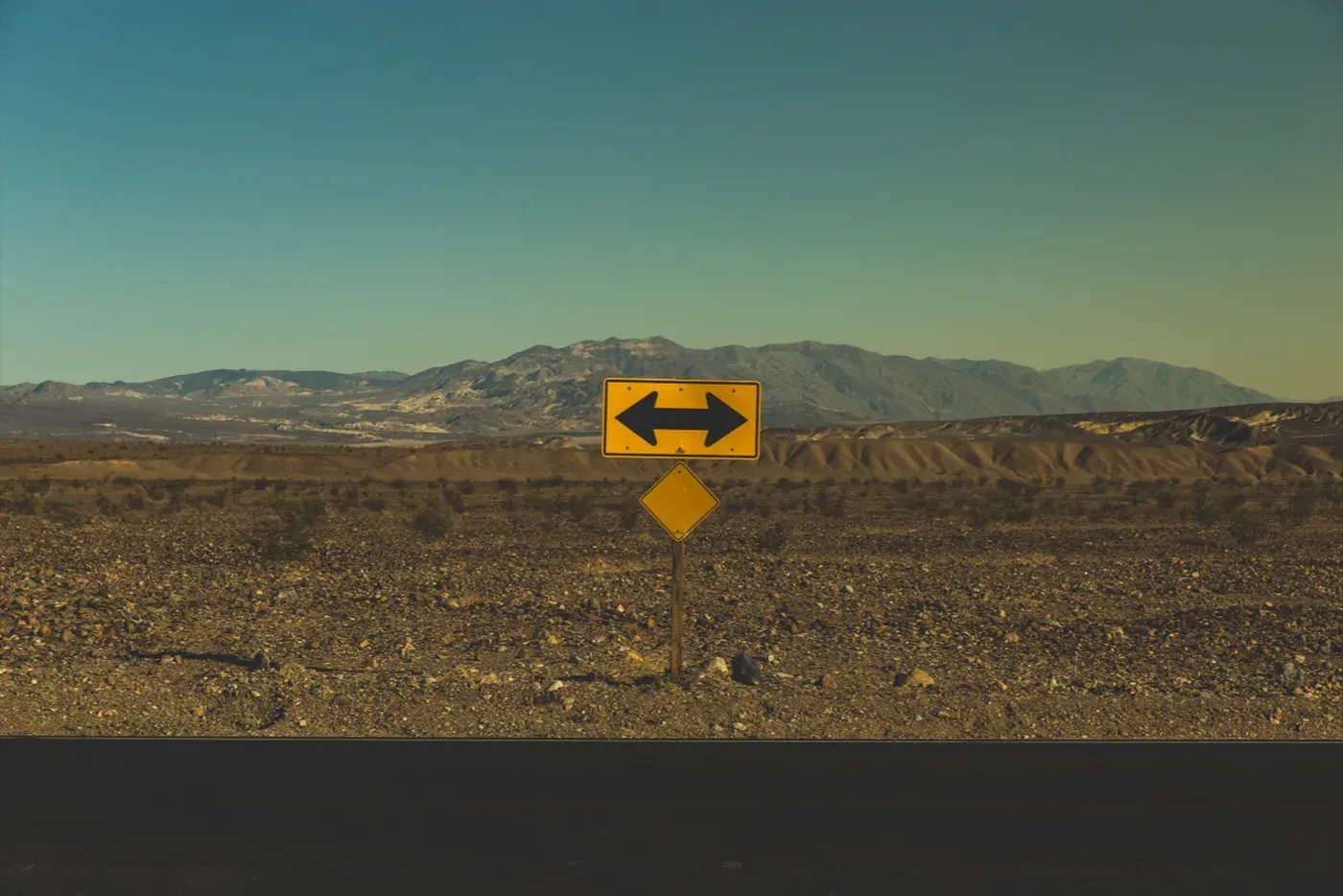 A yellow road sign with a black double-headed arrow pointing left and right, standing in flat desert beside a road with mountains behind.
