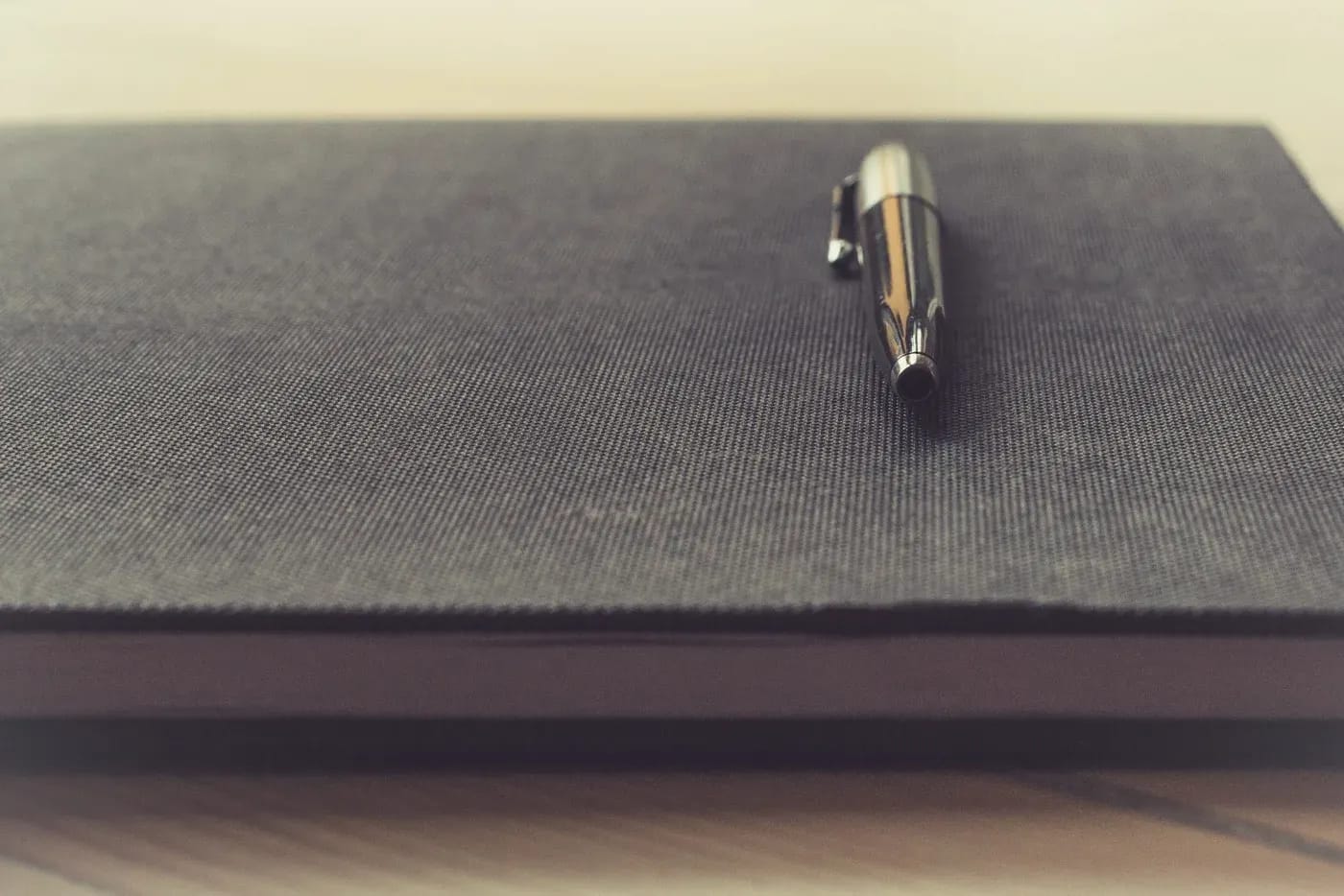 A metal pen resting on a closed dark fabric-covered notebook on a wooden desk.