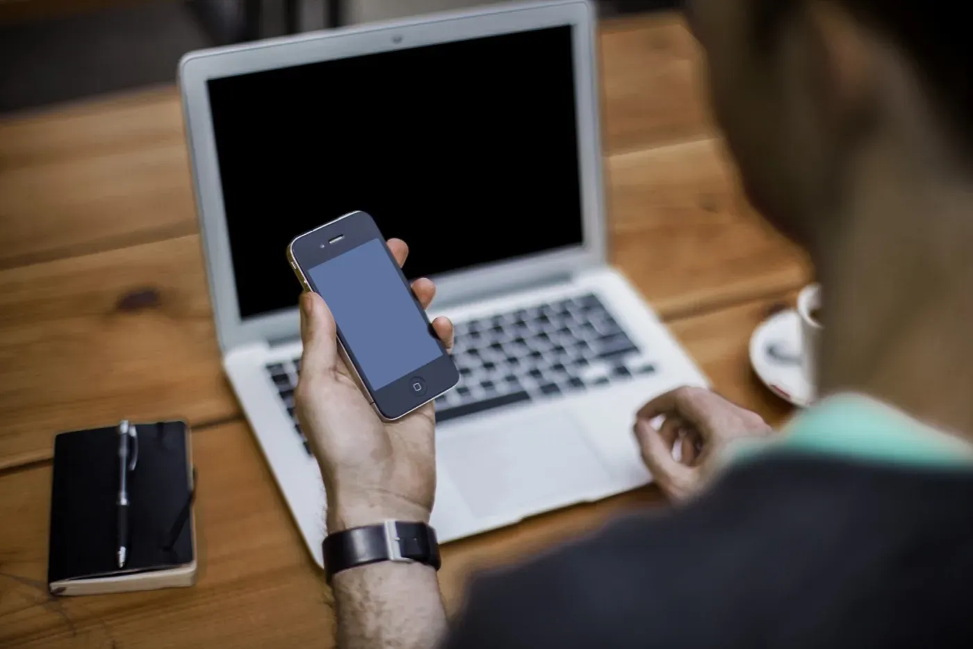 Over-the-shoulder view of a person at a wooden desk holding a smartphone in one hand, with an open laptop, a notebook and pen, and a coffee in front of them.