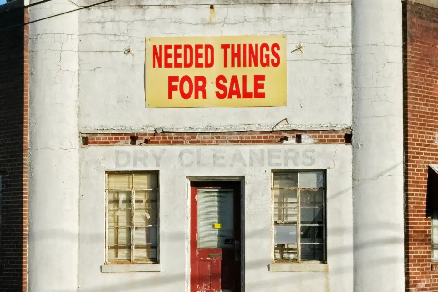 The front of an old white-painted shop with a yellow banner reading Needed Things For Sale above the faded painted words Dry Cleaners, a red door and two shuttered windows.
