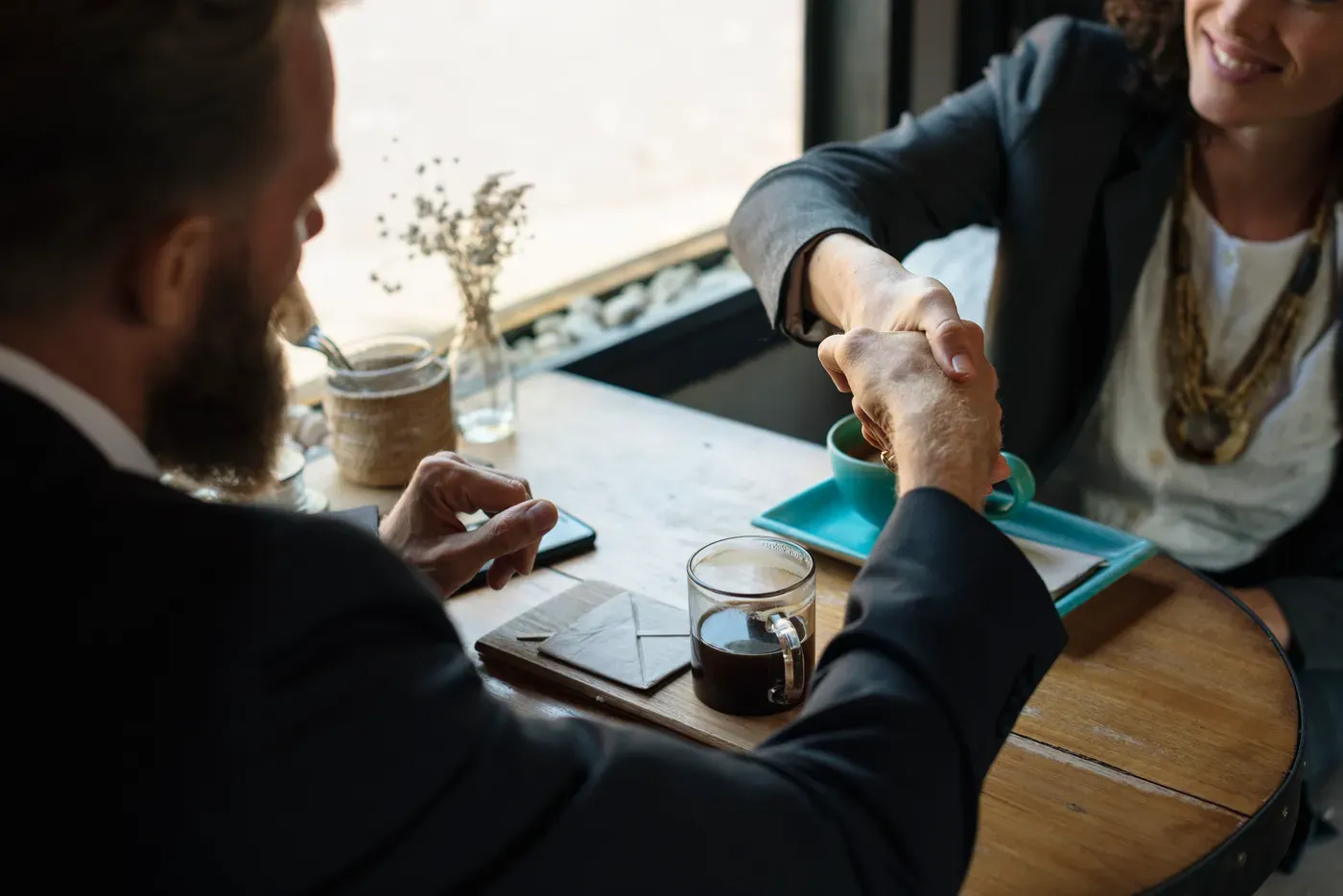 Two people in dark blazers shake hands across a round wooden cafe table by a window, with a glass mug of black coffee, a teal cup and a phone on the table.