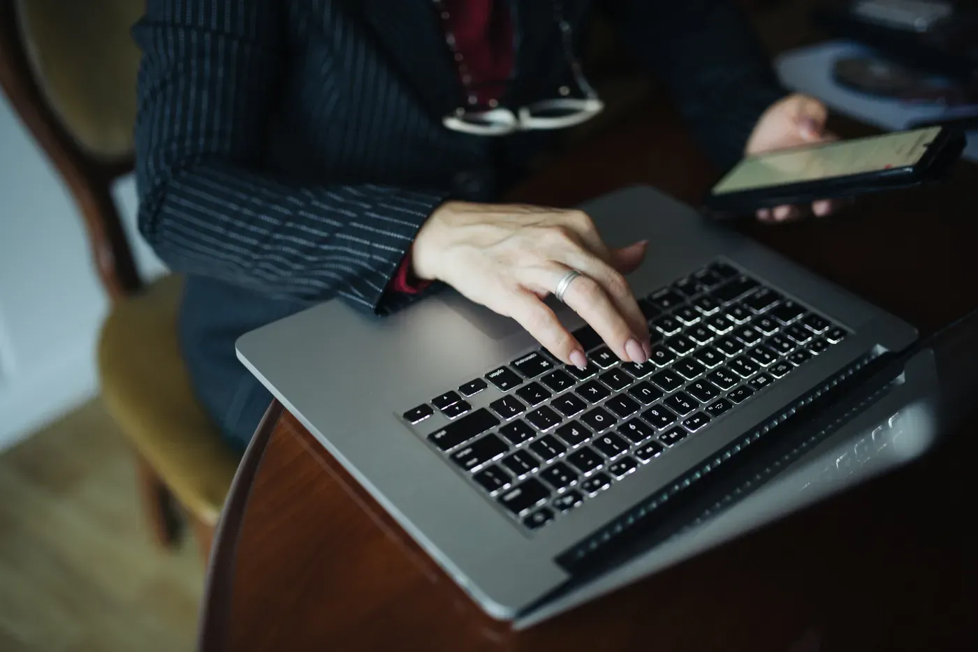 A person in a dark pinstriped jacket types on a silver laptop with a backlit keyboard at a wooden table, holding a smartphone in their other hand.