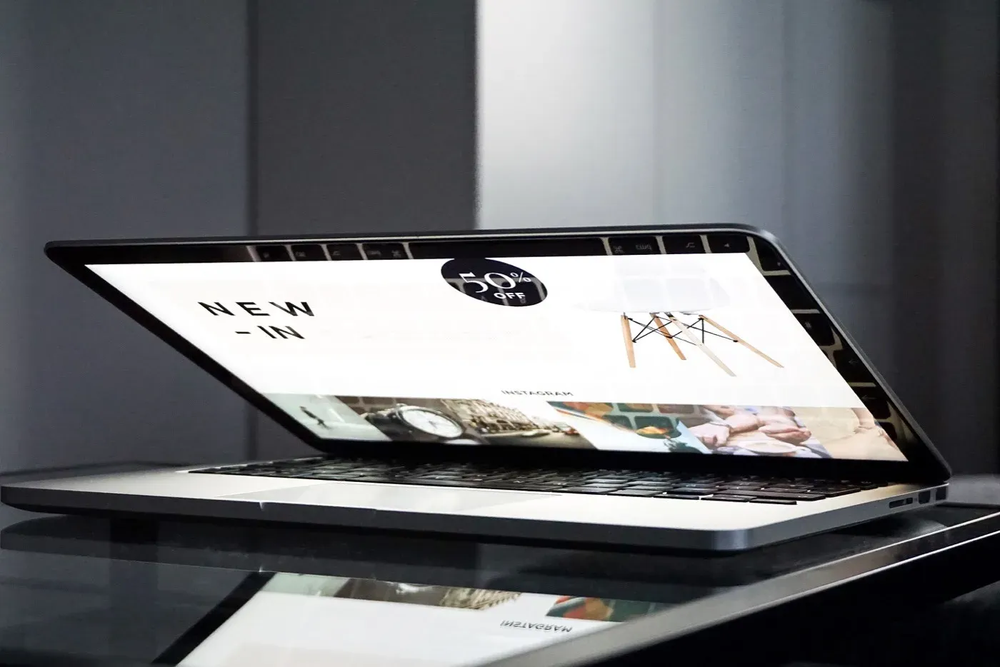 An open laptop on a glossy dark table displaying an online store homepage, with large New In text, product photos and a discount badge on the screen.