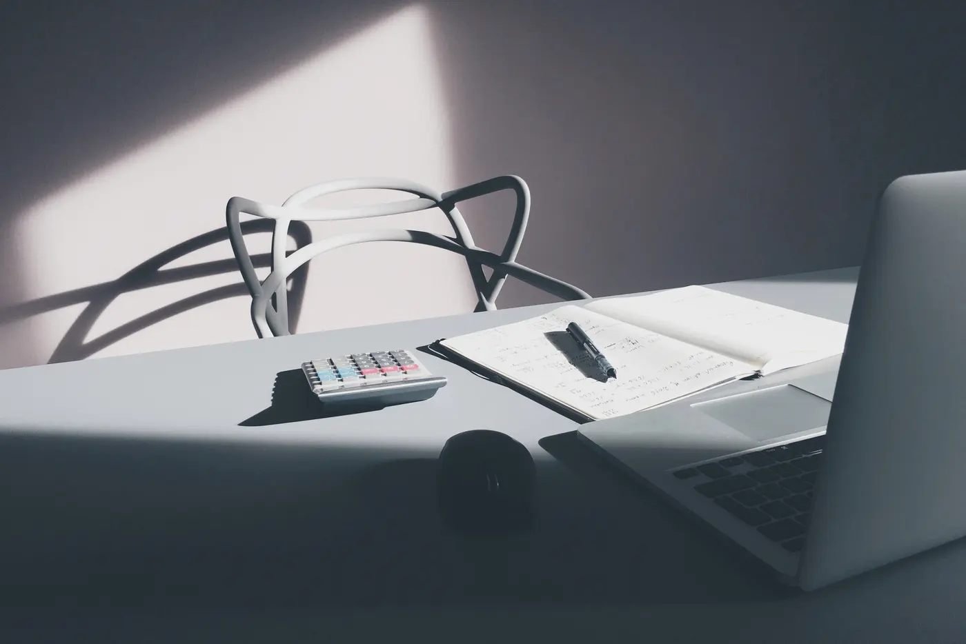 A desk with a white calculator, an open notebook of handwritten figures with a pen, a computer mouse and the edge of an open laptop, beside a white chair in angled sunlight.