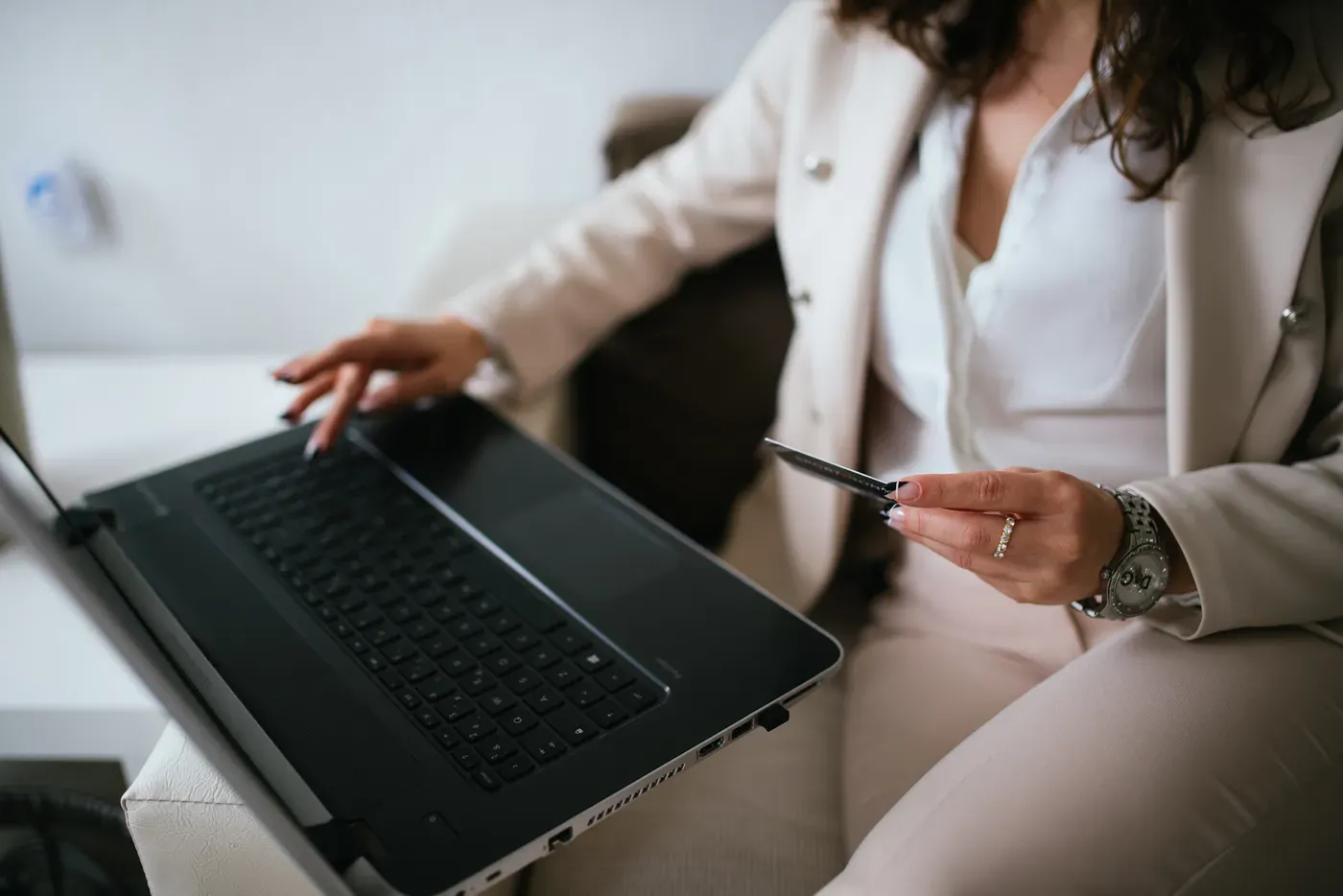 A woman in a cream suit sits with a laptop resting on her lap, typing with one hand and holding a payment card in the other.